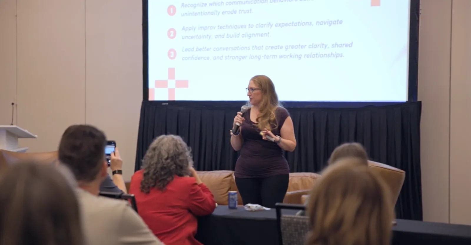 Jennifer Dymond speaking with a microphone on a conference stage in front of a slide listing the session's learning objectives.